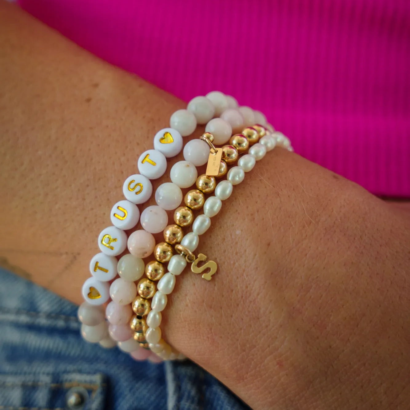A person is wearing multiple bracelets on their wrist, including a white bead bracelet with the word "TRUST" on it, a gold bead bracelet, and a pearl bracelet.