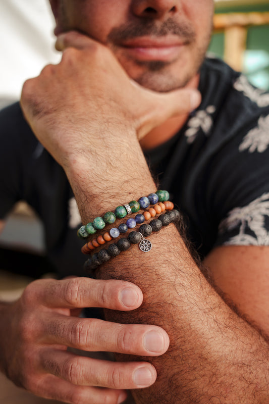 A close-up of a man's hand wearing multiple bracelets, including a green and blue beaded bracelet, a brown wooden bracelet, and a black and orange bead bracelet.